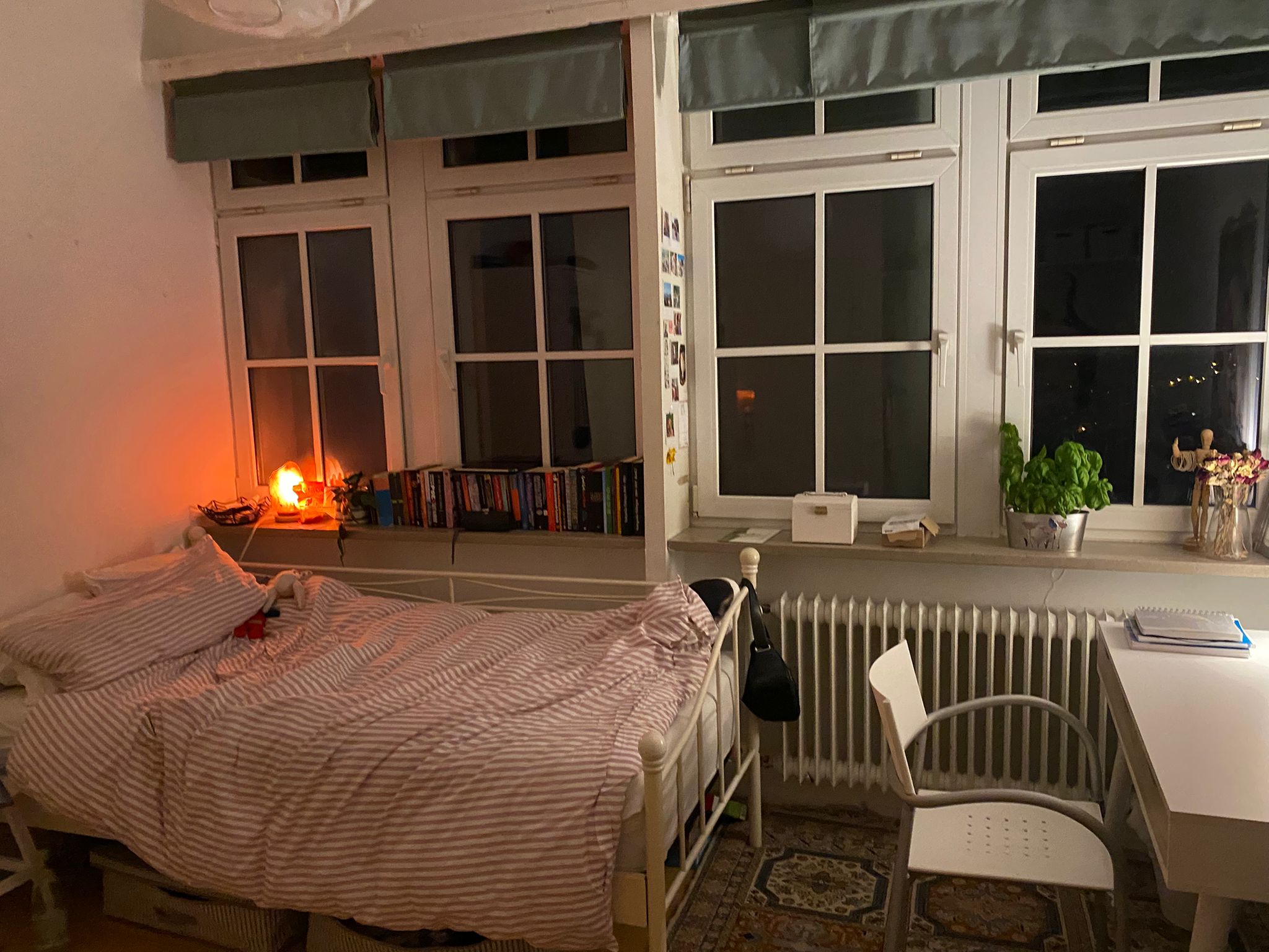 Zimmer mit Bett vor der Fensterfront, Schreibtisch und Stuhl, abends bei warmem Lampenlicht